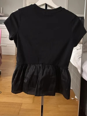 Svart topp med volangkant - Svart topp med korta ärmar och rund hals. Nedre delen har en volangkant i ett glansigt tyg som ger en snygg kontrast till den matta överdelen. Perfekt att styla med jeans eller kjol för en trendig look. Storlek xs