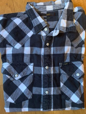 Svartvit rutig skjorta från Lee - Säljer en svartvit rutig skjorta från Lee i slim fit-modell. Skjortan har klassisk krage, tryckknappar framtill och två bröstfickor med lock. Perfekt för dig som gillar en stilren och tidlös look.
