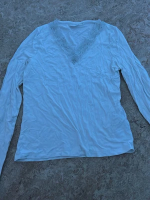 Vit långärmad topp med spets - Säljer en vit långärmad topp med v-ringning och spetsdetalj vid halsen. Toppen har en enkel och stilren design, perfekt att matcha med olika outfits. Materialet känns mjukt och lätt.