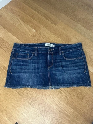 Mörkblå jeanskjol från Abercrombie - Säljer en mörkblå jeanskjol från Abercrombie & Fitch. Kjolen är kort, har rå kant nertill, klassiska fem fickor och bälteshällor. Snygga kontrastsömmar och bakfickor med broderad detalj. Perfekt till en avslappnad look.                                                                Mått: • Midja: 44 cm • Längd: 26 cm • Stussvidd: 47 cm