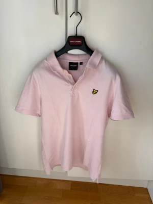 Lyle & Scott: Piké-Tröja - Säljer en ljusrosa pikétröja från Lyle & Scott med klassisk krage och två knappar framtill. Tröjan har korta ärmar och den ikoniska gula loggan broderad på bröstet. Tillverkad i mjuk bomull för en skön känsla. 🎉SlimFit 🎉För mer info skriv dm!
