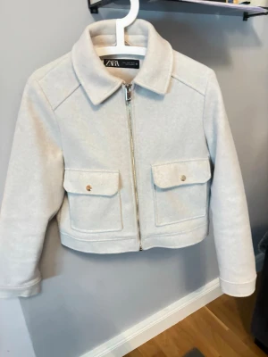 Beige kort jacka från Zara - Stilren beige jacka från Zara i kort modell med rak passform. Jackan har två stora fickor framtill med lock och guldfärgade knappar samt dragkedja. Tillverkad i mjukt, ull-liknande material och har klassisk krage. Perfekt för lager-på-lager och trendiga outfits. Passar även S