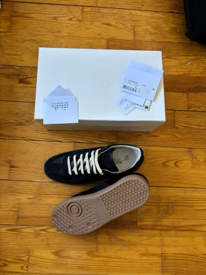 Svarta sneakers från Maison Margiela - Snygga svarta sneakers från Maison Margiela med vita skosnören och beige sula. Skorna har en klassisk låg silhuett och är tillverkade i en mix av mocka och läder. Ikonisk logotyp på plösen och stilren design som passar till det mesta.