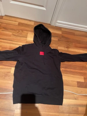 Svart hoodie från Hugo med röd logga - Snygg svart hoodie från Hugo med klassisk röd logga på bröstet. Hoodien har huva med dragsko och en stor magficka framtill. Tillverkad i mjukt bomullsmaterial som känns skönt mot huden. Perfekt för en avslappnad och trendig stil.