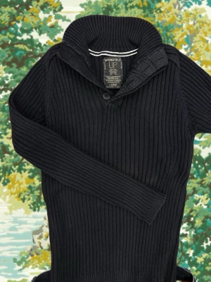 Svart ribbstickad tröja Springfield - Svart ribbstickad tröja från Springfield med hög krage och knappdetalj vid halsen. Tröjan har långa ärmar och en smal passform som ger en clean och stilren look. Perfekt för dig som gillar klassiska plagg med en twist.