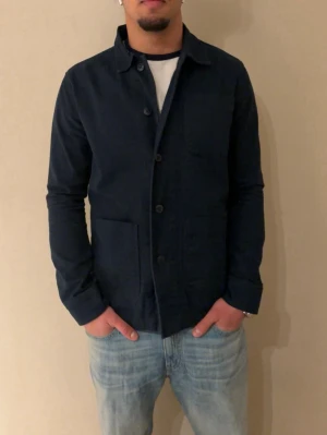 Mörkblå overshirt från Knowledge cotton appereal  - Stilren mörkblå overshirt med klassisk krage och knäppning framtill. Två stora framfickor och en bröstficka ger en avslappnad look. Tillverkad i ett mjukt tyg som känns bekvämt och passar perfekt till jeans. Enkel och tidlös design.