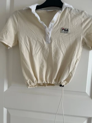 Beige croppad topp från FILA - Säljer en beige croppad topp från FILA med vit krage och korta ärmar. Toppen har en snygg snörning i midjan och en liten logga på bröstet. Perfekt för dig som gillar sportig och avslappnad stil.