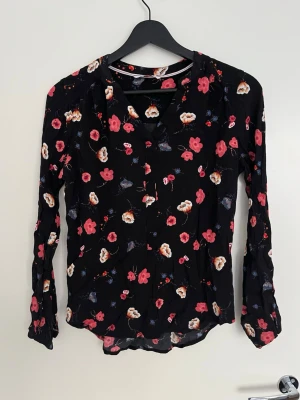 Blommig blus från Tommy Hilfiger - Snygg svart blus från Tommy Hilfiger med färgglatt blommönster i rött, vitt och blått. Blusen har lång ärm och en lätt v-ringad halsringning. Tillverkad i ett tunt, luftigt material som ger en skön känsla och ett avslappnat fall.