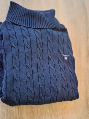 Mörkblå kabelstickad polotröja GANT - Snygg mörkblå kabelstickad polotröja från Gant med broderad logga på bröstet. Tröjan har hög ribbad krage, långa ärmar och ribbade muddar vid ärmslut och nederkant. Perfekt för kyliga dagar och ger en klassisk look. Den har används några få gånger och då har den varit varsamt använd. Priset är inte hugget i sten