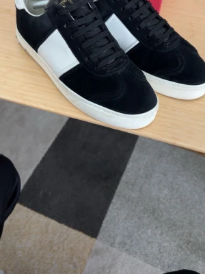 Valentino Garavani svarta sneakers - Snygga svarta sneakers från Valentino Garavani med vita detaljer och vit sula. Skorna har ovandel i mocka och läder, klassisk låg siluett och svarta skosnören. Perfekta för dig som gillar stilrena och exklusiva sneakers.