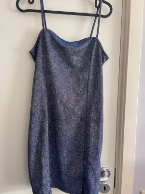 Glittrig blå kort klänning - Snygg glittrig blå kortklänning med tunna axelband och rak passform. Perfekt för dig som vill sticka ut med en enkel men festlig look. Materialet har en skimrande finish som fångar ljuset.