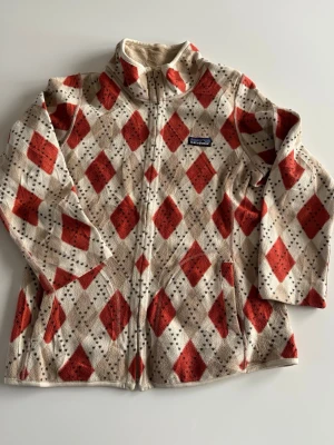 Patagonia, unisex, L  fleecejacka mönstrad  - Säljer en beige och orange fleecejacka från Patagonia med rutigt mönster och dragkedja. Jackan  är mjukt, mysigt material som passar perfekt för vänliga dagar. Klassisk Patagonia-logga på bröstet.