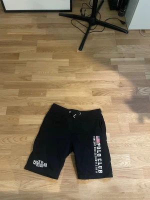 Svarta shorts från Polo Club - Säljer ett par svarta shorts från Polo Club med vita tryck på ena benet och amerikansk flagga. Shortsen har dragsko i midjan och är tillverkade i mjukt material, perfekta för en avslappnad stil.