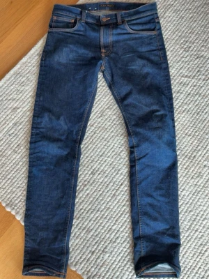 Mörkblå slim jeans från Nudie - Snygga mörkblå slim jeans från Nudie med klassiska orange sömmar och fem fickor. Jeansen har en slim passform och är tillverkade i stretchigt denim för extra komfort. Nudie-logga på bakfickan och patch i läder baktill i midjan.