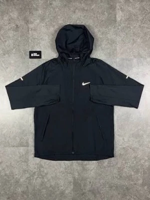 Svart vindjacka med huva från Nike - Snygg svart vindjacka från Nike med hel dragkedja och huva. Jackan har vita Nike-loggor på bröstet och ärmarna, samt två sidofickor. Tillverkad i lätt och tåligt material som passar perfekt för blåsiga dagar.