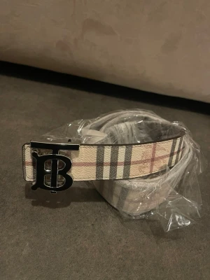 Beige rutigt Burberry bälte - Snyggt bälte från Burberry med klassiskt beige, svart och rött rutmönster. Bältet har en stor metallspänne med Burberrys ikoniska TB-logga i svart. Perfekt accessoar för att lyfta din outfit.