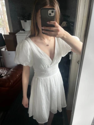 Vit klänning - Superfin vit kortklänning med omlott-design och stora puffärmar. Klänningen har en v-ringad fram och bak samt en stor rosett baktill. Materialet är lätt och plisserat, vilket ger en luftig känsla. Perfekt för sommarens alla tillfällen. Stl 34, passar mig som är xs. Ny pris 699kr säljer för 500kr