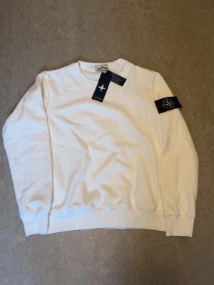 Vit sweatshirt från Stone Island - Snygg vit sweatshirt från Stone Island som är helt oanvänd. Den har en klassisk rund hals och ikonisk svart patch med gult kompassmärke på ärmen, kommer även med ett lila märke som snabbt går att byta ut. Perfekt för dig som vill ha en clean look. Kontakta mig gärna om du har några flera frågor om tröjan. Tröjan skickas ut samma dag eller dagen efter som du har beställt📦