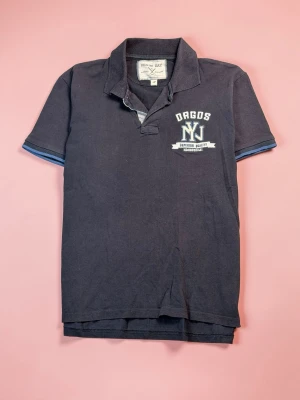 Mörkblå pikétröja från Benny Day - Klassisk mörkblå pikétröja från Benny Day med vit broderad text och emblem på bröstet. Tröjan har korta ärmar, krage och knappslå framtill. Perfekt för en avslappnad och sportig look.