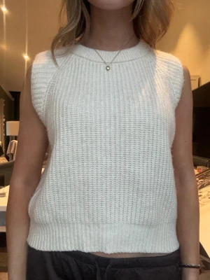 Vit stickad ärmlös tröja - Säljer en vit stickad ärmlös tröja i mjukt material. Tröjan har rund halsringning och ribbade kanter nedtill och vid ärmhålen. Perfekt att styla över en skjorta eller bära som den är för en trendig look.