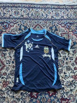 Argentina Adidas fotbollströja S - Mörkblå Argentina fotbollströja från Adidas med ljusblå och vita detaljer, nummer 19 och AFA-emblem på bröstet. Tröjan har Stüssy-logga och är tillverkad i lätt, ventilerande material. Perfekt för fotboll eller sportiga outfits.