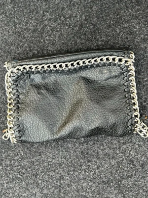 Svart kuvertväska med kedjedetaljer - Snygg svart kuvertväska i fejkläder med grov struktur och coola silverfärgade kedjedetaljer runt kanterna. Väskan har en rektangulär form och stängs med dragkedja upptill. Perfekt accessoar för att lyfta din outfit.