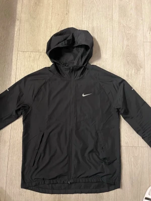 Nike Miller (S) - Snygg svart vindjacka från Nike med dragkedja framtill och justerbar huva. Jackan har två sidofickor med dragkedja och en vit Nike-logga på bröstet. Tillverkad i lätt och vindtätt material, perfekt för blåsiga dagar.