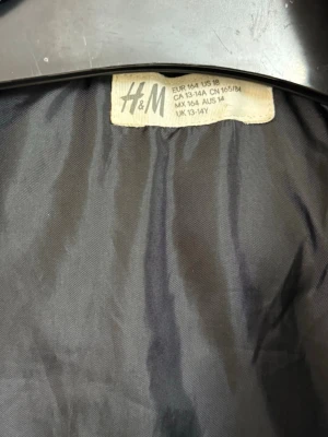 Svart pufferjacka från H&M - Snygg svart pufferjacka från H&M med huva och breda quiltade paneler. Jackan har en cool detalj med ett svartvitt band med text på huvan. Perfekt för kalla dagar och riktigt skön att bära.