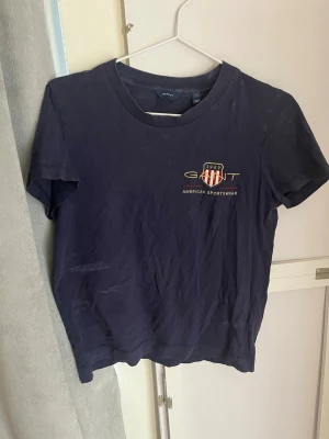 Mörkblå t-shirt från Gant - Snygg mörkblå t-shirt från Gant med tryckt logga och emblem på bröstet. Klassisk rund halsringning och korta ärmar. Tillverkad i mjuk bomull som känns skön mot huden. Perfekt till jeans eller shorts för en avslappnad look. Har ett antal defekter som syns på bild 2, 3 och 5