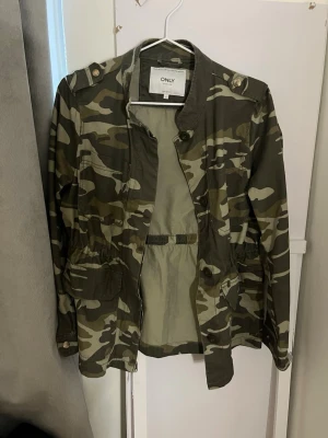 Camouflage parkajacka från ONLY - Snygg camouflagefärgad parkajacka från ONLY med dragsko i midjan och knappar på axlarna. Jackan har flera fickor framtill och är tillverkad i bomullsmix. Perfekt för dig som vill ha en streetstyle-look.