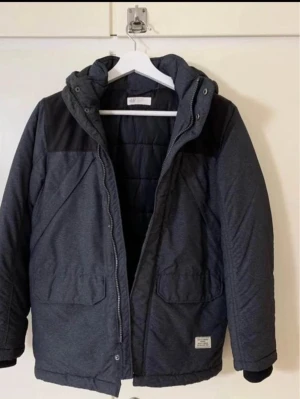 Mörkgrå dunjacka från H&M - Snygg mörkgrå dunjacka från H&M med svarta detaljer över axlarna och huva. Jackan har två stora fickor framtill, dragkedja och tryckknappar. Perfekt för kalla vinterdagar och har en stilren look.