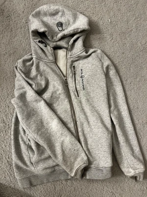 Grå hoodie från Sail Racing - Snygg grå hoodie från Sail Racing med dragkedja och huva. Har en bröstficka med dragkedja och logga på bröstet samt på huvan. Tillverkad i mjuk bomullsblandning, perfekt för chill dagar. Klassisk passform och ribbade muddar.