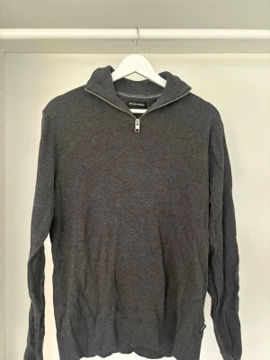 Mörkgrå halvzip tröja Jack & Jones - Mörkgrå stickad tröja från Jack & Jones med halvzip och hög krage. Tröjan har långa ärmar och ribbade muddar vid ärmslut och nederkant. Perfekt för lager på lager och enkel att matcha med olika stilar.