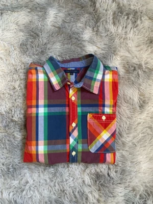 Flerfärgad rutig skjorta från TommyHilfiger  - Säljer en färgglad rutig skjorta från TommyHilfiger med lång ärm och klassisk krage. Skjortan har knappar framtill och en bröstficka med matchande rutmönster. Tillverkad i mjuk bomull och har inslag av rött, blått, grönt och gult. Den är i storlek 164 och priset kan diskuteras vid snabb affär 