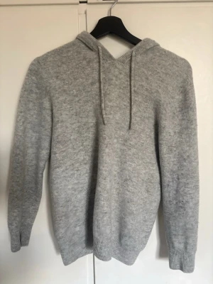 Ljusgrå Kashmir hoodie - Mysig ljusgrå stickad hoodie med huva och dragsnören. Tröjan har en avslappnad passform och ribbade muddar vid ärmslut och nederkant. Perfekt för en chill och stilren look. Materialet känns mjukt och skönt mot huden.