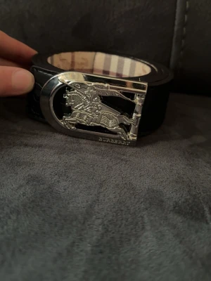  Burberry bälte  - Snyggt svart skärp från Burberry med klassiskt rutmönster och präglad logga längs bandet. Spännet är i silverfärgad metall med en ikonisk riddare. Perfekt accessoar för att lyfta din outfit.