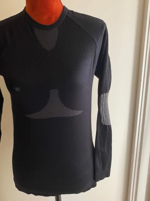 Svart funktionsundertröja med mönster - Säljer en svart långärmad funktionsundertröja med grått meshmönster på ryggen och armbågarna. Tröjan är tight i modellen och har rund hals. Perfekt för träning eller som baslager vid utomhusaktiviteter.