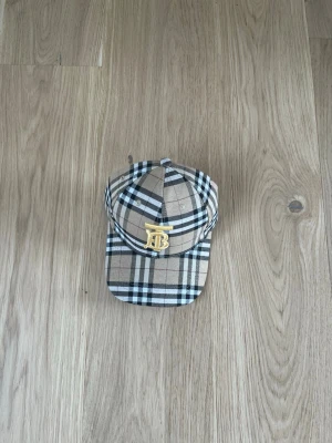 Beige rutig Burberry keps - Säljer en klassisk beige rutig keps från Burberry med det ikoniska rutmönstret och ett guldigt TB-monogram framtill. Kepsen har böjd skärm och justerbar rem bak. Insidan är fodrad med Burberry-loggor. Perfekt för dig som gillar streetstyle och lyxiga detaljer.