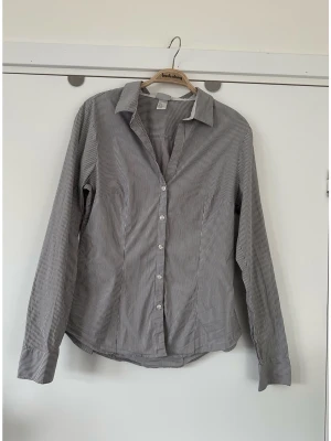 H&M - grå vit randig skjorta - storlek 42 - Stilren figursydd grå-vit-randig skjorta / blus från H&M i storlek 42.  Figurnära modell med långa ärmar och knäppning framtill. Krage och manschetter med knappar. Passar bra för en klassisk och stilren look. 74% bomull, så bra material som andas. Mycket gott skick, som ny!