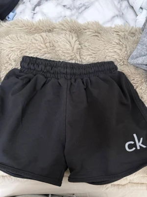 Svarta shorts från Calvin Klein - Svarta shorts från Calvin Klein med elastisk midja och loggan 'ck' tryckt på ena benet. De har en avslappnad passform och är perfekta för sommarens alla aktiviteter. Tillverkade i mjuk bomull för maximal komfort.
