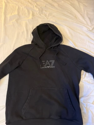 Svart EA7 hoodie från Emporio Armani - Svart hoodie från Emporio Armani EA7 med  luva och ficka framtill. Logga tryckt på bröstet och band med Armani-tryck längs ärmarna. Storlek M men passar S. Pris kan diskuteras.