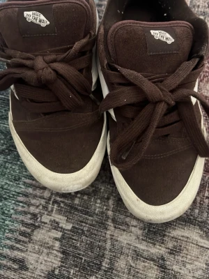 Bruna Vans sneakers med vit sula - Säljer ett par bruna Vans sneakers med klassisk vit sula och bruna skosnören. Skorna har den ikoniska 'Off The Wall'-loggan på plösen och är tillverkade i canvas och mockaliknande material. Perfekta för dig som gillar streetwear och skate-stil.