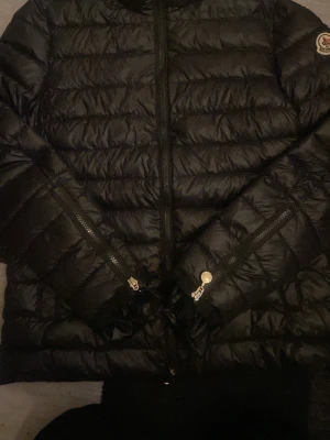 Svart dunjacka från Moncler - Snygg svart dunjacka från Moncler med quiltad design och två dragkedjeförsedda fickor framtill. Jackan har hög krage och klassisk Moncler-logga på ärmen. Perfekt för kalla vinterdagar och riktigt stilren look.