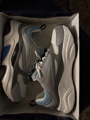 Dior vita och grå sneakers - Chunky sneakers från Dior i vitt, grått och ljusblått. Skorna har en modern siluett med meshpaneler och detaljer i läder. Tjock sula och snörning framtill. Perfekta för dig som gillar streetwear och exklusiva märken.