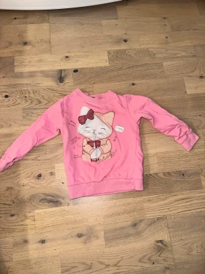 Rosa sweatshirt med kattmotiv - Söt rosa sweatshirt från KiggaLilly med ett gulligt kattmotiv framtill. Tröjan har ribbade muddar vid ärmslut och nederkant samt en rund halsringning. Perfekt för dig som gillar djurmotiv och vill ha en mysig och lekfull stil.