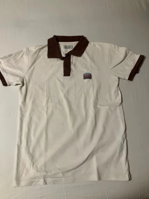 Vit piké med bruna detaljer - Snygg vit pikétröja från Jeans Dept med brunt krage och bruna muddar vid ärmarna. Tröjan har knappslå med två knappar och en broderad logga på bröstet. Perfekt för en avslappnad men stilren look.