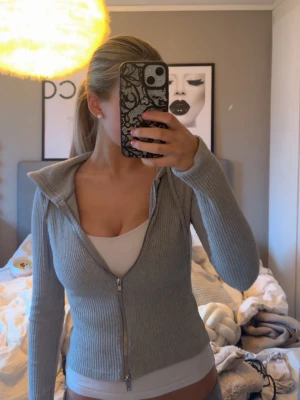 Grå ribbad hoodie med dragkedja - Säljer en grå ribbad hoodie med dragkedja framtill. Hoodien har långa ärmar, åtsittande passform och en huva. Materialet är stretchigt och mjukt, perfekt för en avslappnad och trendig look.