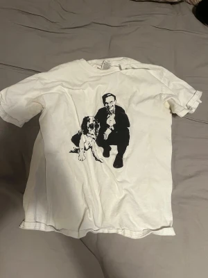 Vit t-shirt med tryck av man och hund - Knappt använd Vit t-shirt med svart tryck av en man i kostym som sitter bredvid en hund. Klassisk passform och rund halsringning. Perfekt för dig som gillar unika och grafiska motiv.
