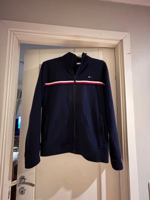 Marinblå zip hoodie Tommy Hilfiger - Snygg marinblå hoodie från Tommy Hilfiger med dragkedja framtill och diskret logga på bröstet. Röd och vit rand går tvärs över bröstet för en sportig look. Tillverkad i mjukt bomullsmaterial och har två fickor fram. Perfekt för en avslappnad stil.