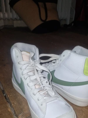 Nike vita high-top sneakers med grönt swoosh - Säljer ett par vita high-top sneakers från Nike med grönt swoosh på sidorna och gul detalj på hälen. Skorna har klassisk snörning och är tillverkade i läder med textilfoder. Perfekta för dig som vill ha en retro och sportig look. Storlek 36,5 nästan aldrig använt kanske 1-2 gånger. 